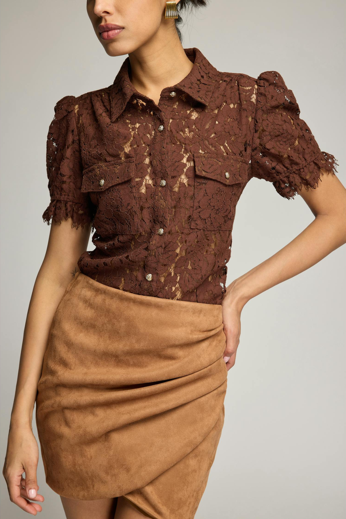 A model wearing a brown lace short-sleeve button-up shirt with puff sleeves and a tan draped faux-suede mini skirt, photographed against a light gray studio background.