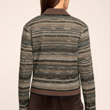 A woman viewed from the back wearing a cropped knit cardigan with horizontal textured stripes in olive green, beige, and brown, paired with dark brown pants, against a solid light beige background.