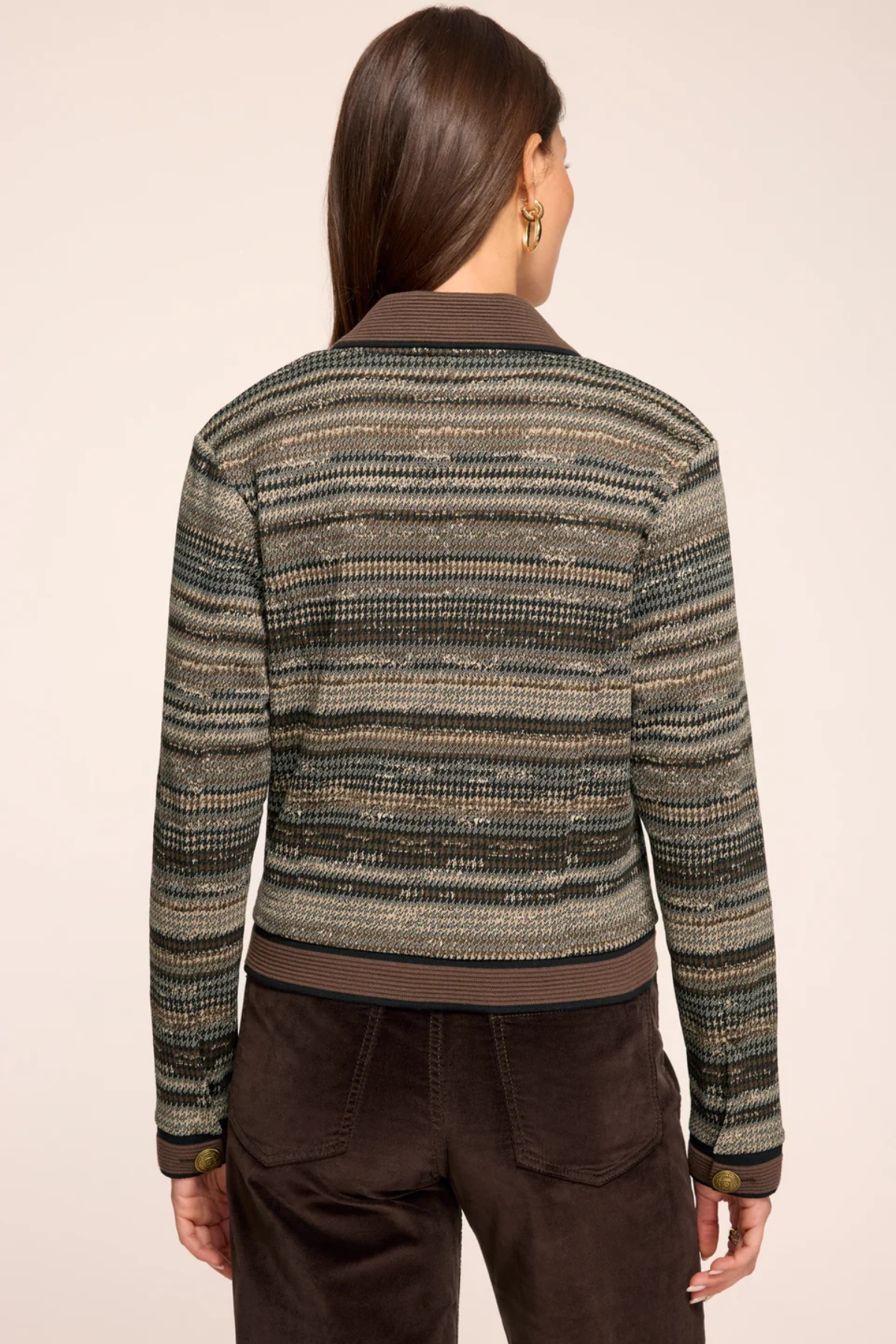 A woman viewed from the back wearing a cropped knit cardigan with horizontal textured stripes in olive green, beige, and brown, paired with dark brown pants, against a solid light beige background.