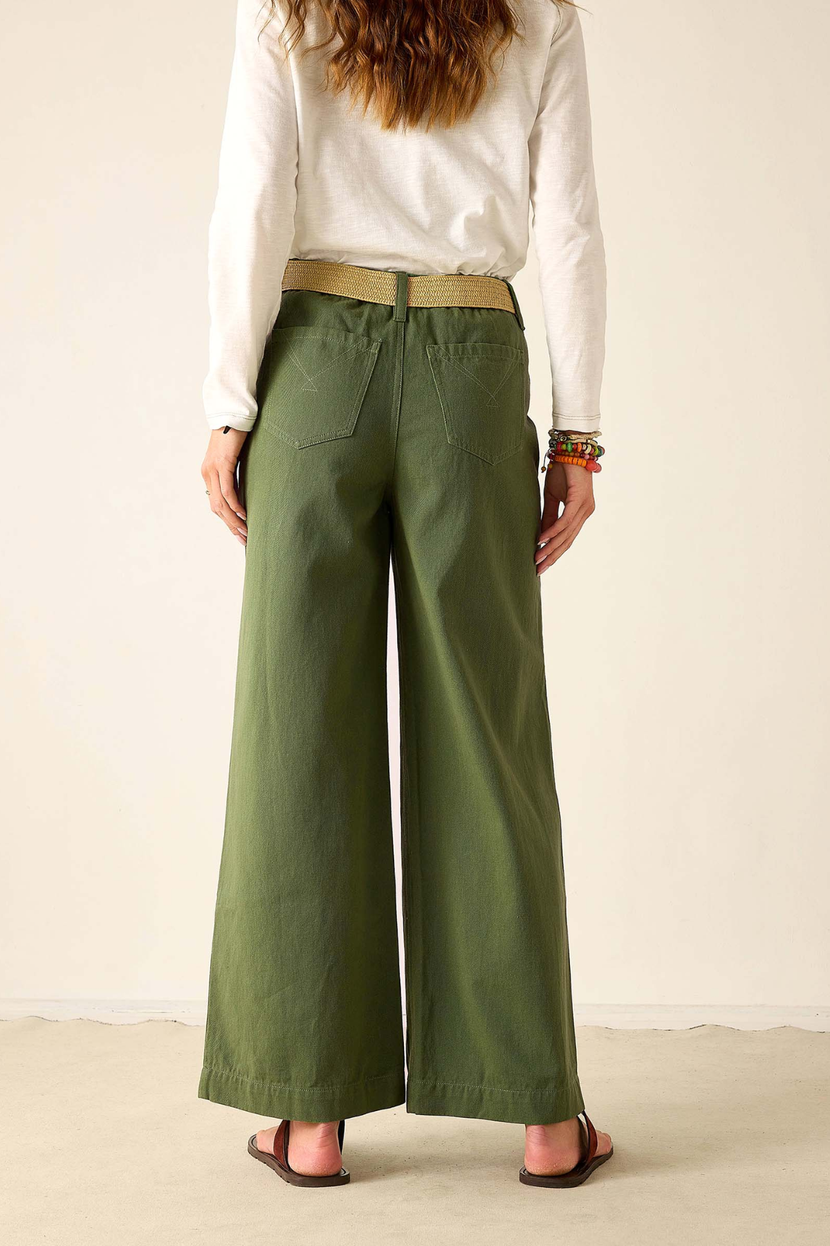 A person shown from the back wearing olive green wide-leg pants with a tan belt, a white long-sleeve top, and brown sandals against a plain cream studio background.