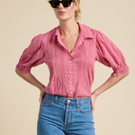 A woman modeling a pink and red vertically striped short-sleeve blouse with puff sleeves and blue jeans against a solid beige background.