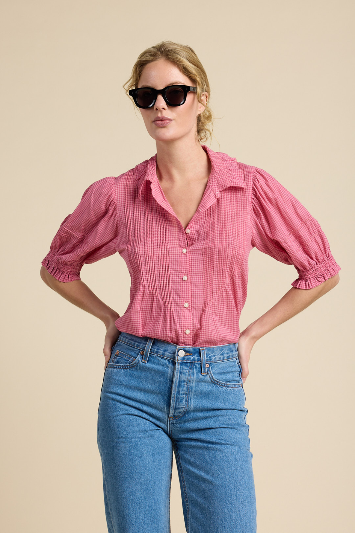 A woman modeling a pink and red vertically striped short-sleeve blouse with puff sleeves and blue jeans against a solid beige background.