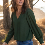 A woman wearing a dark green long-sleeve blouse with puffed shoulders and blue jeans, standing outdoors in a wooded area with warm sunlight filtering through the trees.