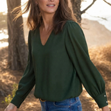 A woman wearing a dark green long-sleeve blouse with puffed shoulders and blue jeans, standing outdoors in a wooded area with warm sunlight filtering through the trees.