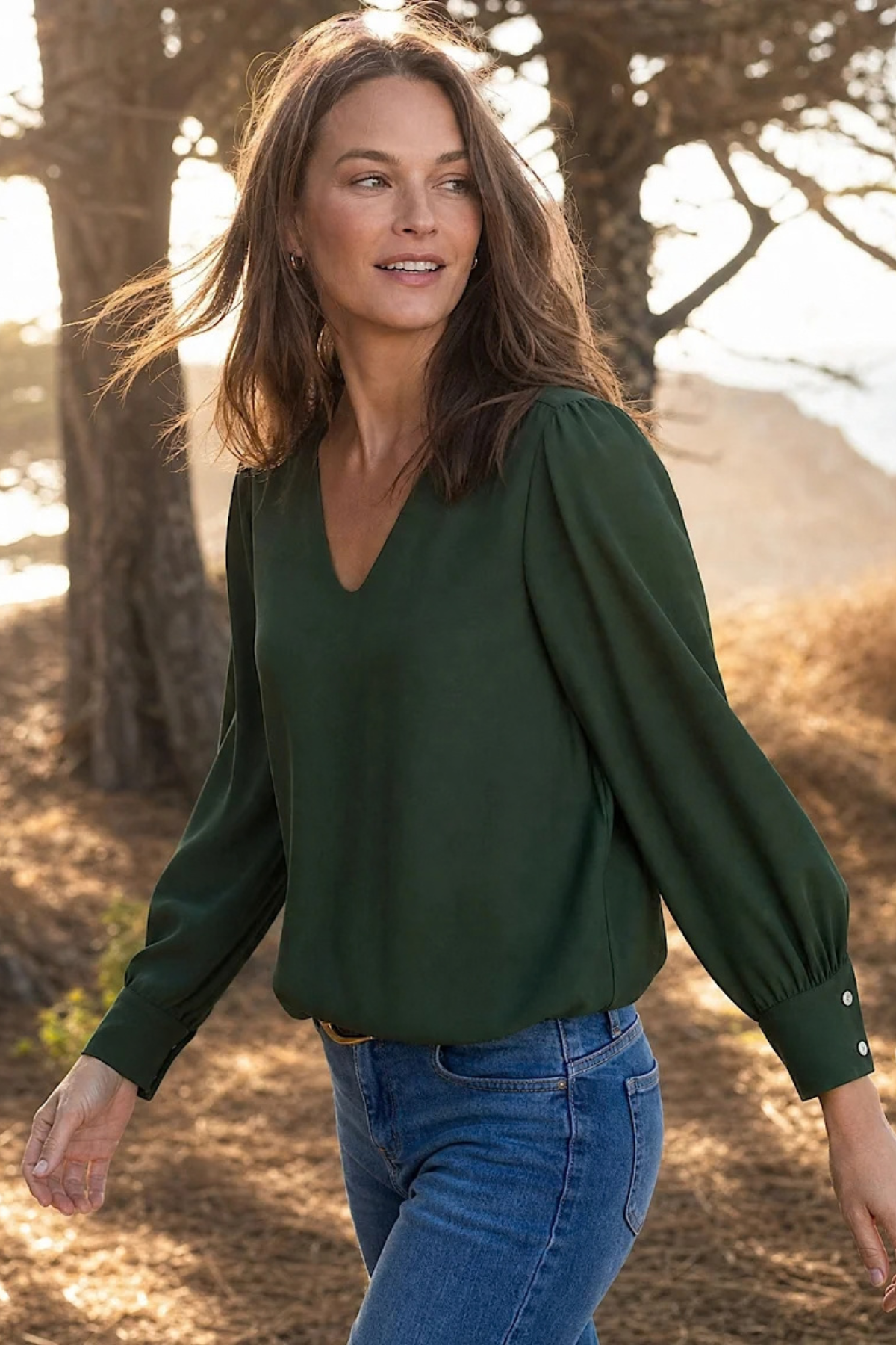 A woman wearing a dark green long-sleeve blouse with puffed shoulders and blue jeans, standing outdoors in a wooded area with warm sunlight filtering through the trees.