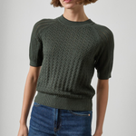 A woman wearing a dark olive green short-sleeve knit sweater with a textured cable pattern, paired with blue jeans, photographed against a neutral light gray background.