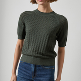 A woman wearing a dark olive green short-sleeve knit sweater with a textured cable pattern, paired with blue jeans, photographed against a neutral light gray background.