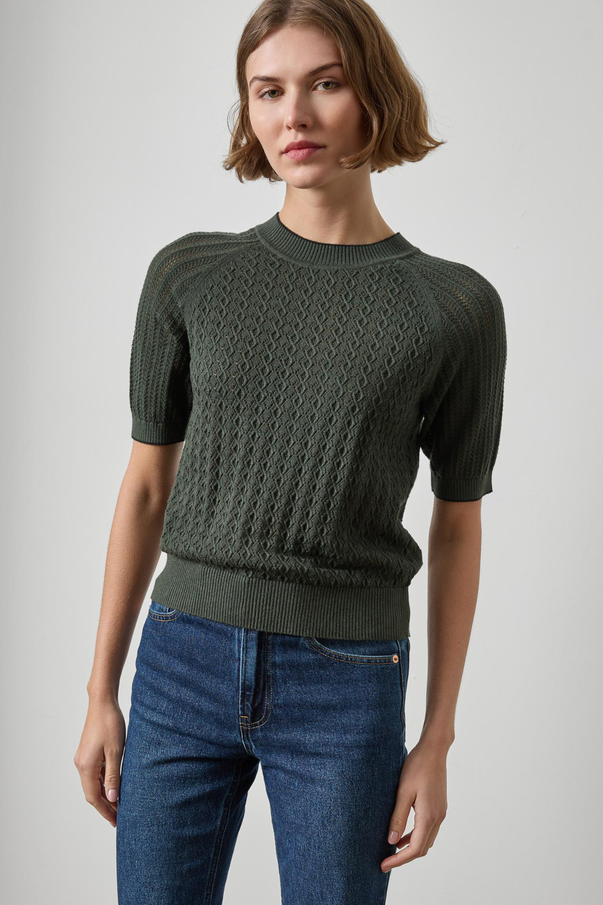 A woman wearing a dark olive green short-sleeve knit sweater with a textured cable pattern, paired with blue jeans, photographed against a neutral light gray background.