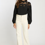 Woman wearing a black blouse with floral lace details and white pants on a white background