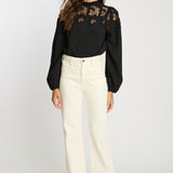 Woman wearing a black blouse with floral lace details and white pants on a white background