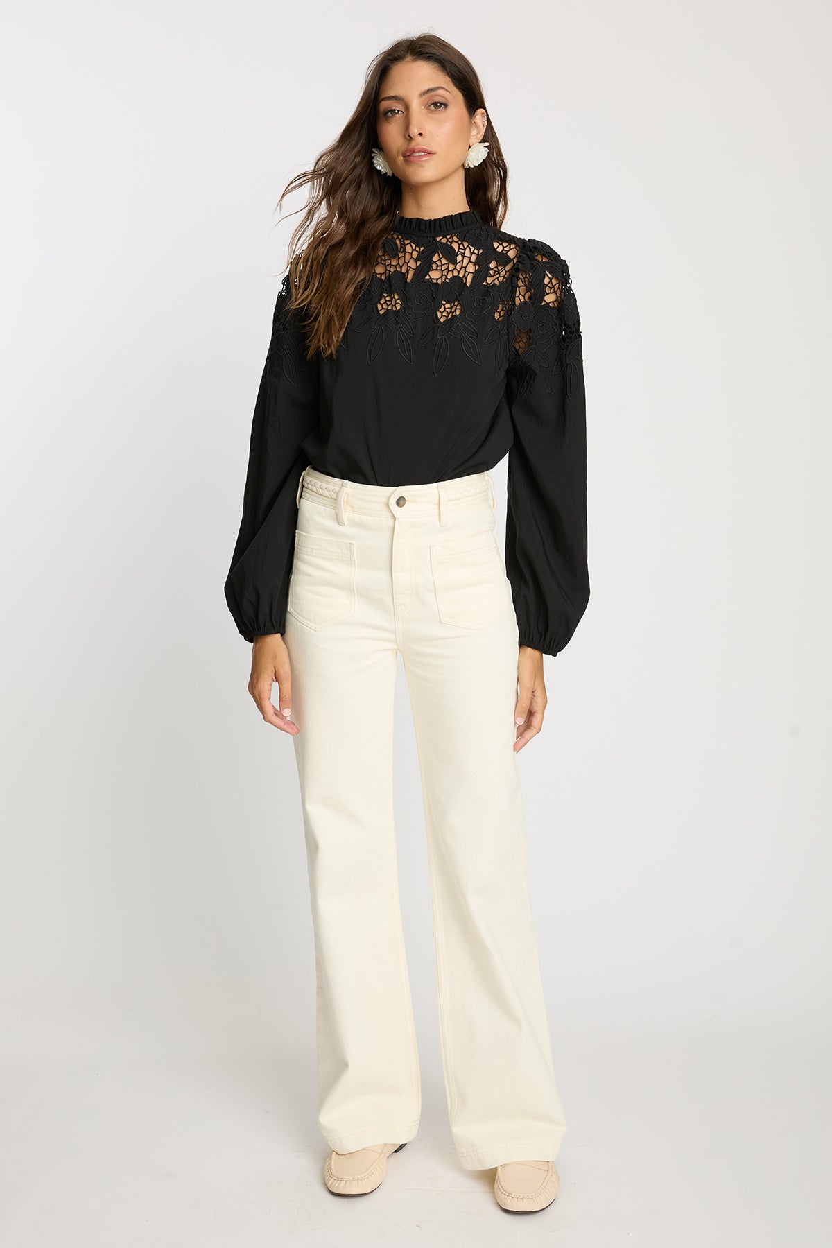 Woman wearing a black blouse with floral lace details and white pants on a white background