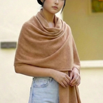 A woman wearing a soft camel-colored knit wrap or shawl draped over her shoulders, paired with light blue jeans, standing against a warm-toned beige wall with a hanging light fixture in the background.
