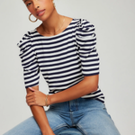 Woman wearing a striped shirt and jeans sitting on a stool against a plain background