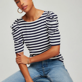 Woman wearing a striped shirt and jeans sitting on a stool against a plain background