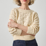 Woman wearing a beige knitted sweater and dark jeans against a plain background