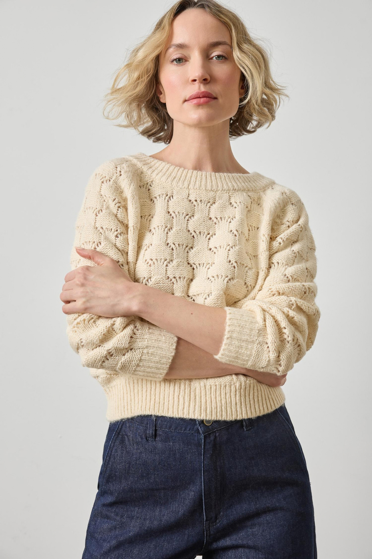 Woman wearing a beige knitted sweater and dark jeans against a plain background