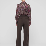 A model shown from the back wearing a long-sleeve high-neck blouse with a purple and pink paisley pattern and dark brown wide-leg trousers, set against a plain light gray studio background.