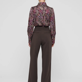 A model shown from the back wearing a long-sleeve high-neck blouse with a purple and pink paisley pattern and dark brown wide-leg trousers, set against a plain light gray studio background.