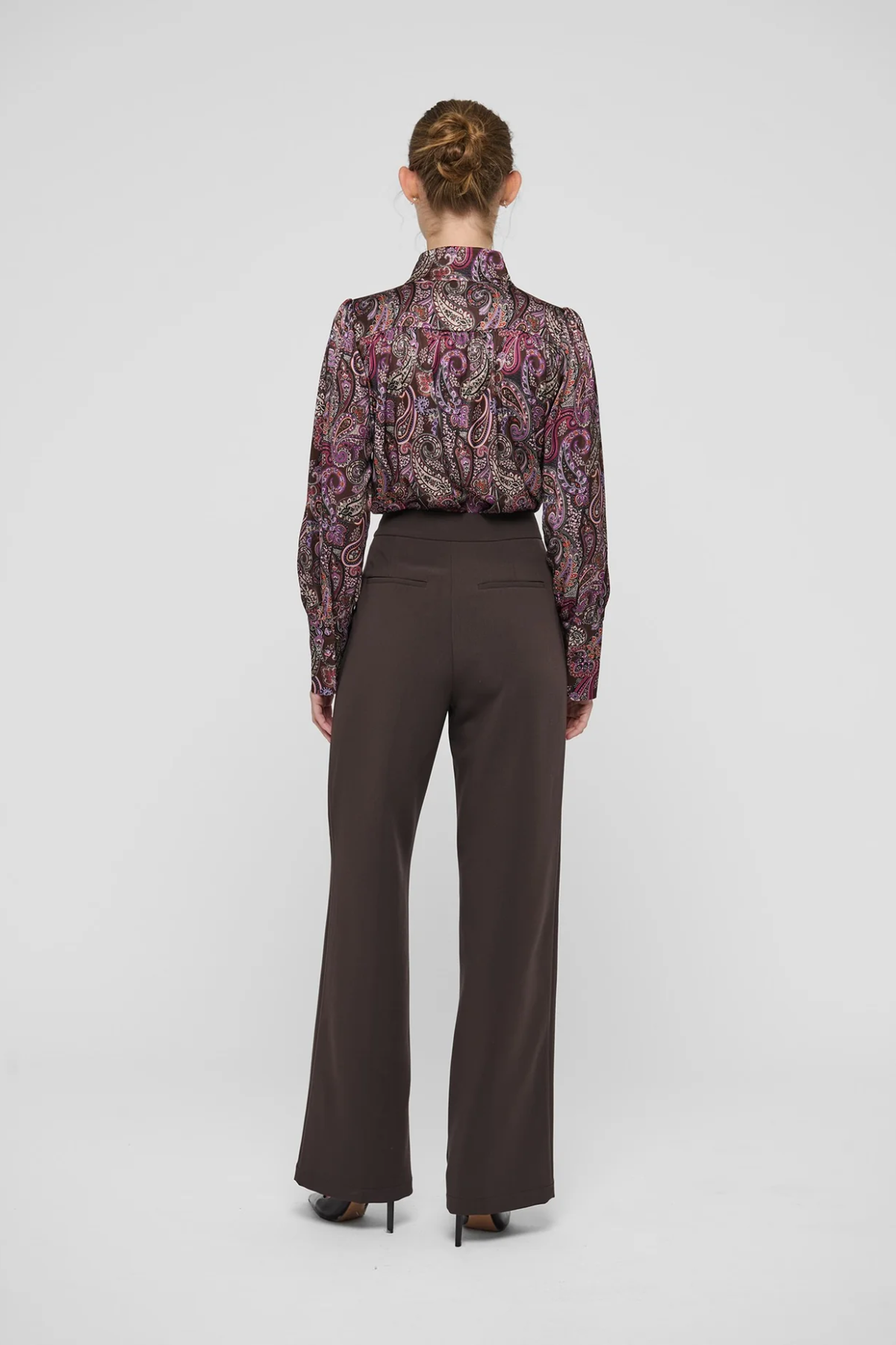 A model shown from the back wearing a long-sleeve high-neck blouse with a purple and pink paisley pattern and dark brown wide-leg trousers, set against a plain light gray studio background.