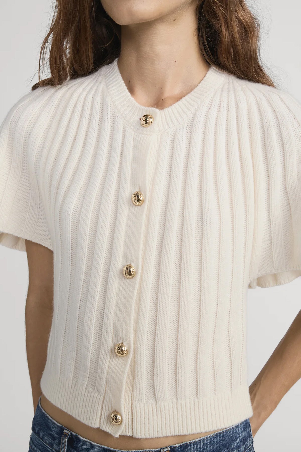 White ribbed knit cardigan with gold buttons worn by a person on a white background