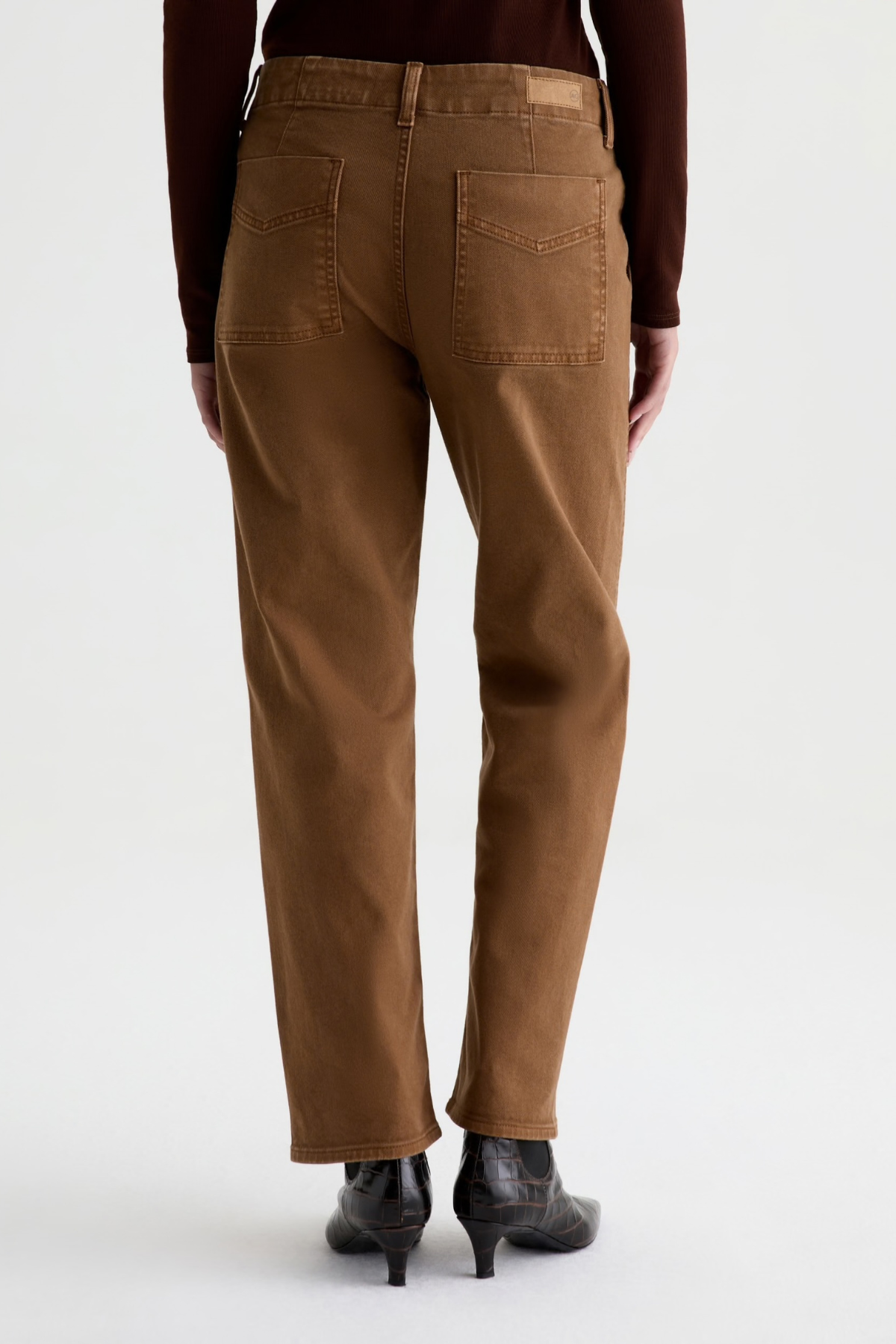 A person shown from the back wearing brown straight-leg jeans, a dark brown long-sleeve top, and black ankle boots against a light gray studio background.