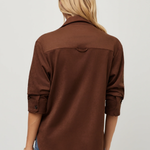 A woman viewed from the back wearing a brown long-sleeve button-up shirt and blue jeans against a plain light gray studio background.