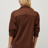 A woman viewed from the back wearing a brown long-sleeve button-up shirt and blue jeans against a plain light gray studio background.