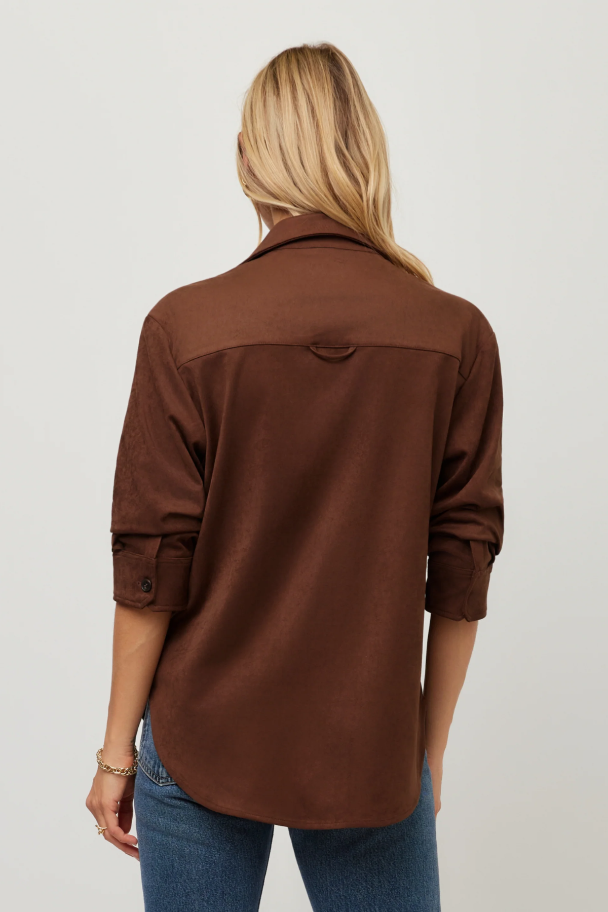 A woman viewed from the back wearing a brown long-sleeve button-up shirt and blue jeans against a plain light gray studio background.