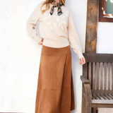 A woman stands indoors wearing a cream knit sweater, a long brown midi skirt, and brown shoes, with a patterned neck scarf. She is posed beside a wooden bench against a white wall with tiled flooring.