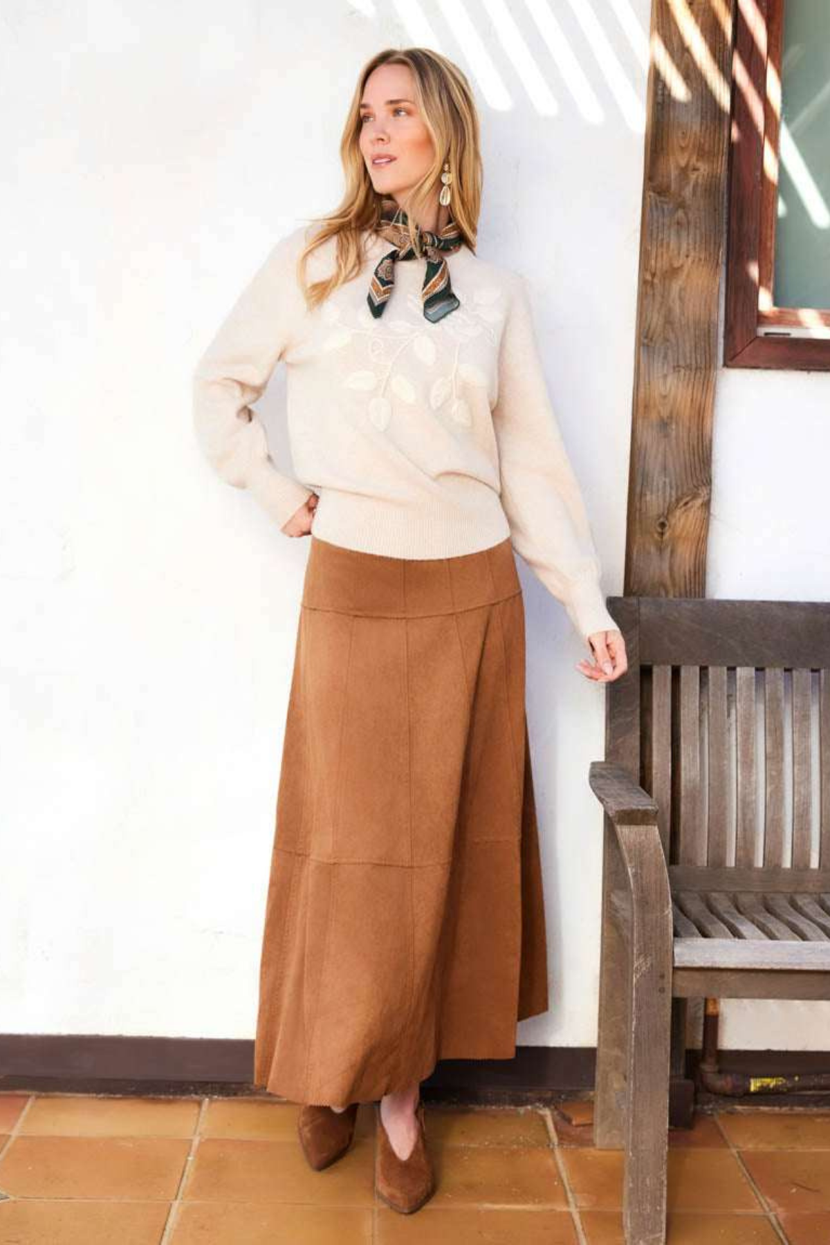 A woman stands indoors wearing a cream knit sweater, a long brown midi skirt, and brown shoes, with a patterned neck scarf. She is posed beside a wooden bench against a white wall with tiled flooring.