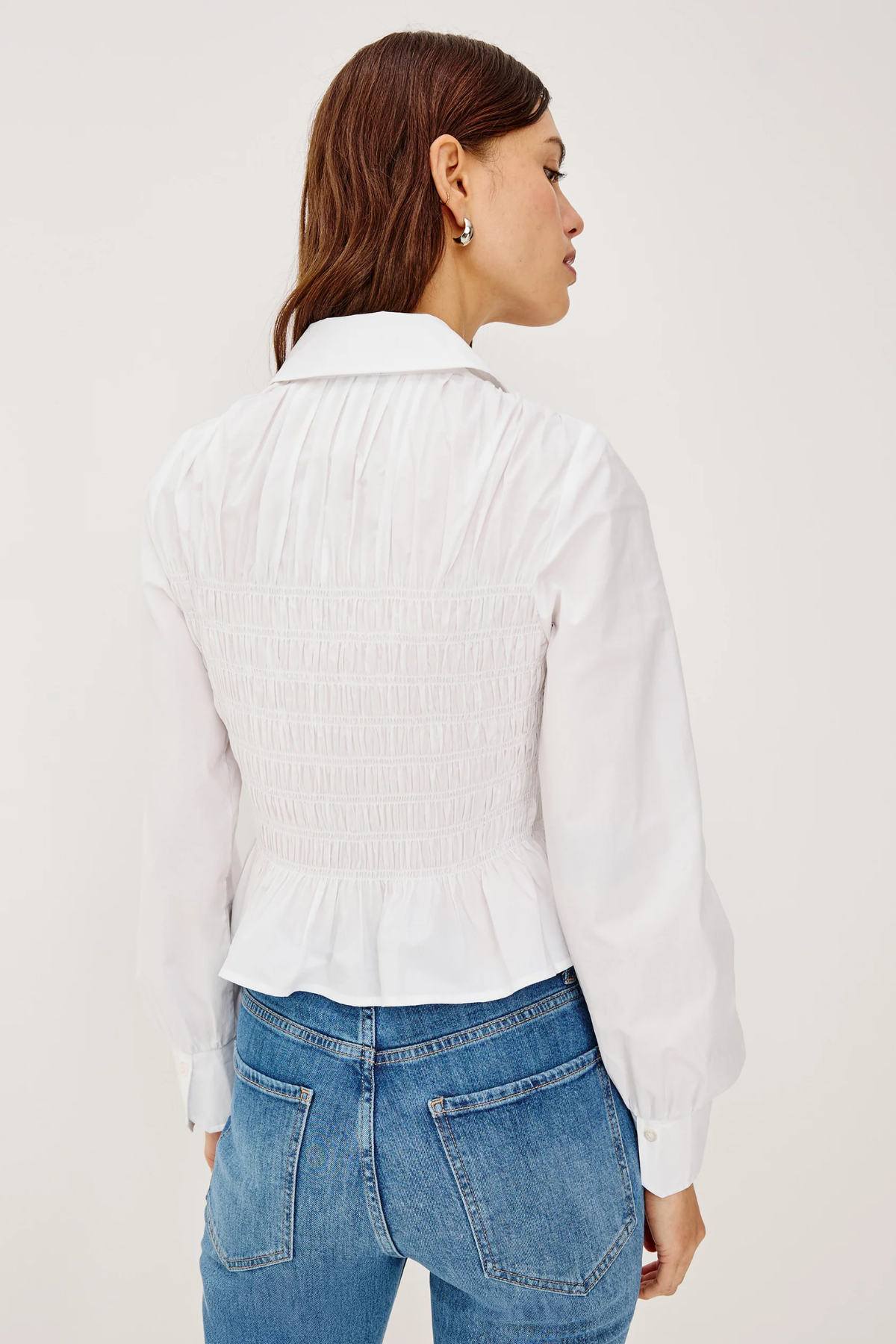 Person wearing a white ribbed sweater over a collared shirt with blue jeans on a light gray background
