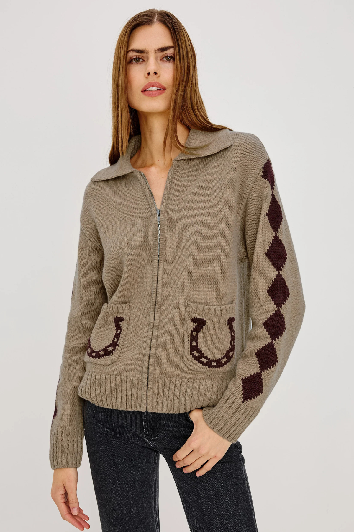 A woman modeling a taupe knit zip-up cardigan with dark brown horseshoe and argyle pattern details, paired with dark jeans against a light gray studio background.