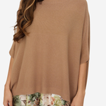 A woman wearing a loose, short-sleeve knit top in a warm camel brown color, layered over a floral print skirt. The top is photographed against a clean, light off-white background.