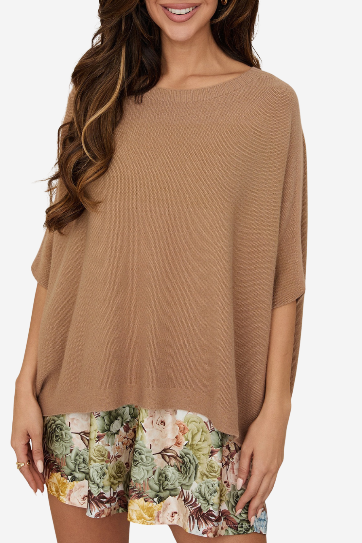 A woman wearing a loose, short-sleeve knit top in a warm camel brown color, layered over a floral print skirt. The top is photographed against a clean, light off-white background.