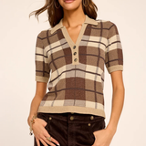 A woman modeling a short-sleeve knit polo-style top with a brown, beige, and cream plaid pattern, paired with dark brown pants, against a plain light beige studio background.