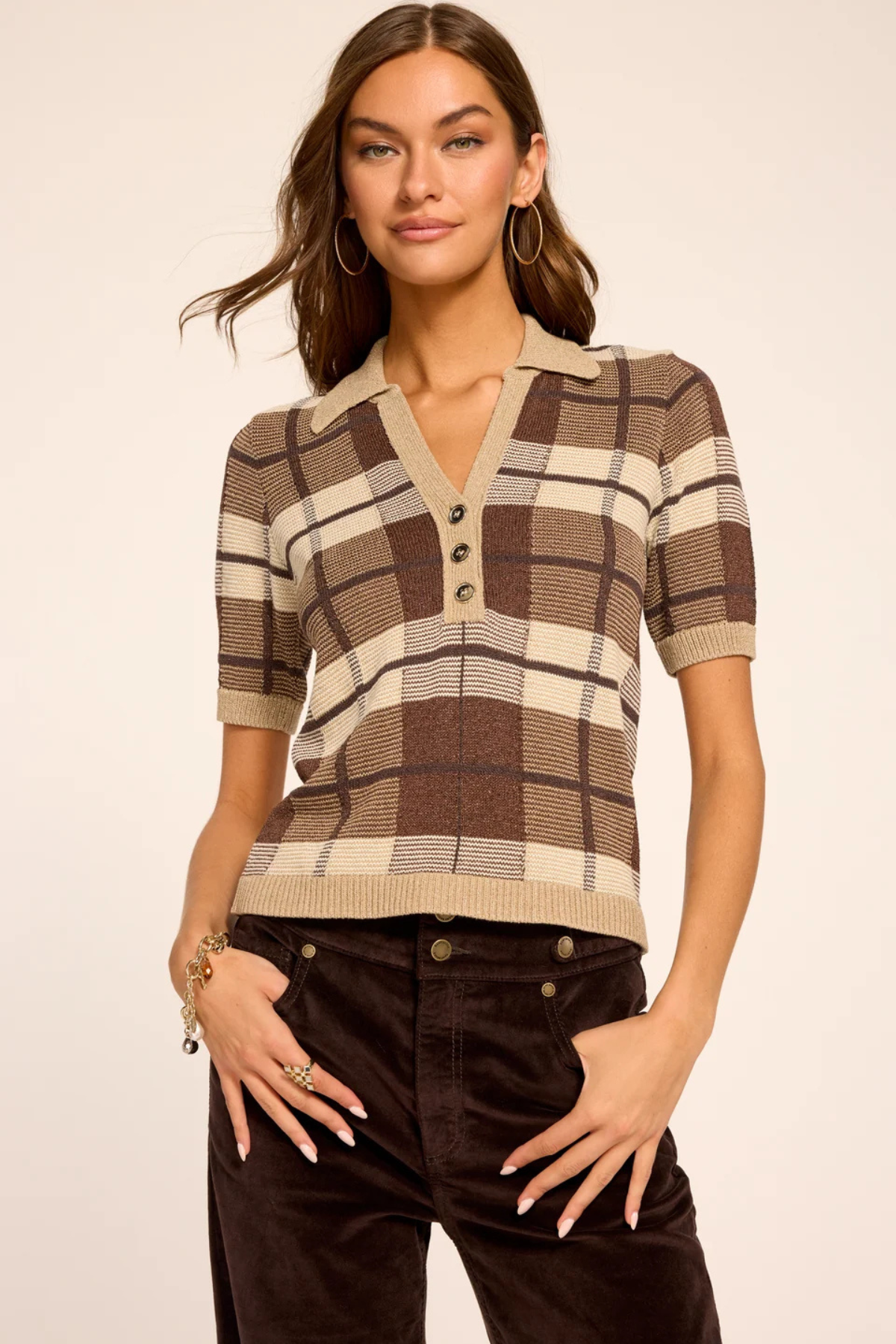 A woman modeling a short-sleeve knit polo-style top with a brown, beige, and cream plaid pattern, paired with dark brown pants, against a plain light beige studio background.