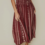 A woman wearing a high-waisted, pleated maxi skirt in burgundy with vertical cream patterns, paired with a white blouse and brown block-heel sandals, set against a plain beige studio background.