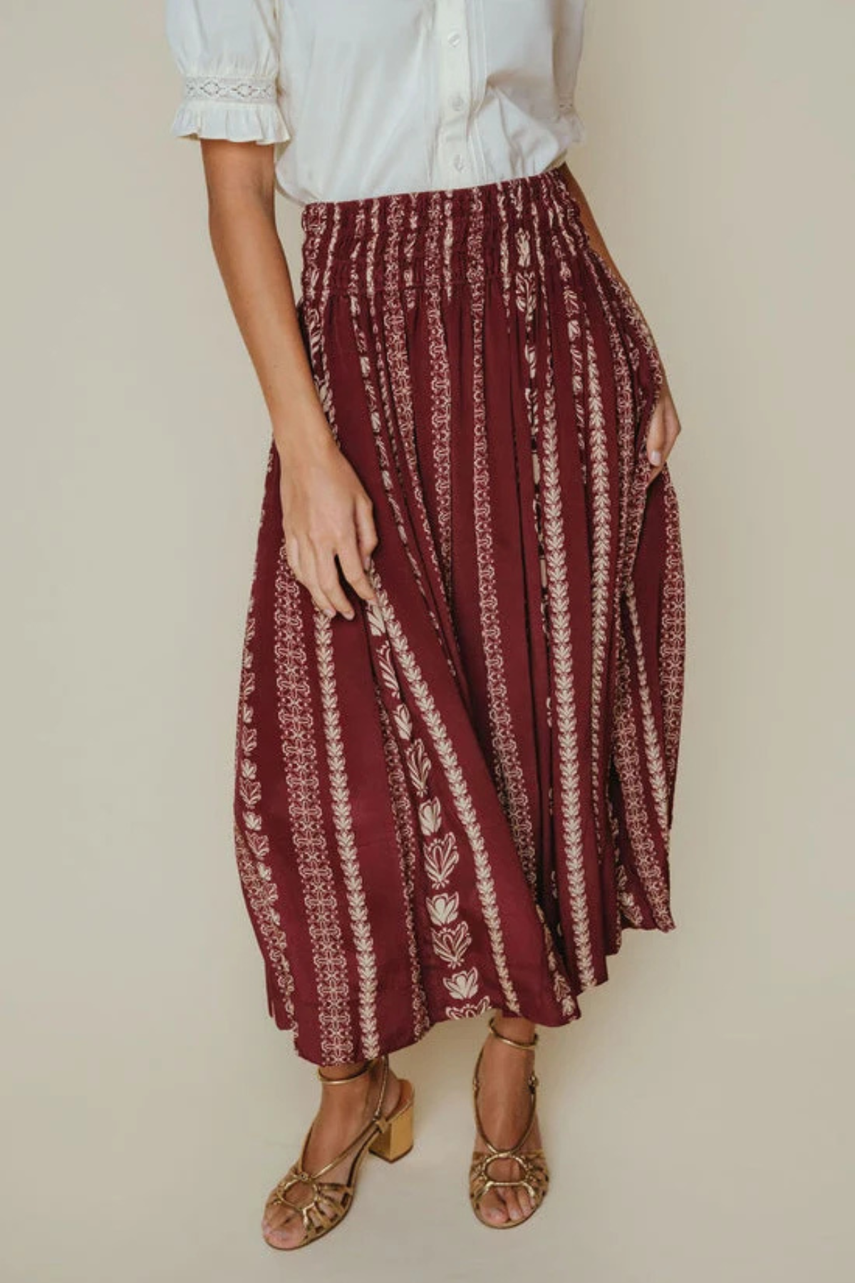 A woman wearing a high-waisted, pleated maxi skirt in burgundy with vertical cream patterns, paired with a white blouse and brown block-heel sandals, set against a plain beige studio background.