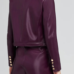 A model shown from the back wearing a matching deep purple faux-leather cropped blazer and shorts set, featuring gold-tone decorative buttons on the cuffs, against a light gray studio background.
