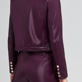 A model shown from the back wearing a matching deep purple faux-leather cropped blazer and shorts set, featuring gold-tone decorative buttons on the cuffs, against a light gray studio background.