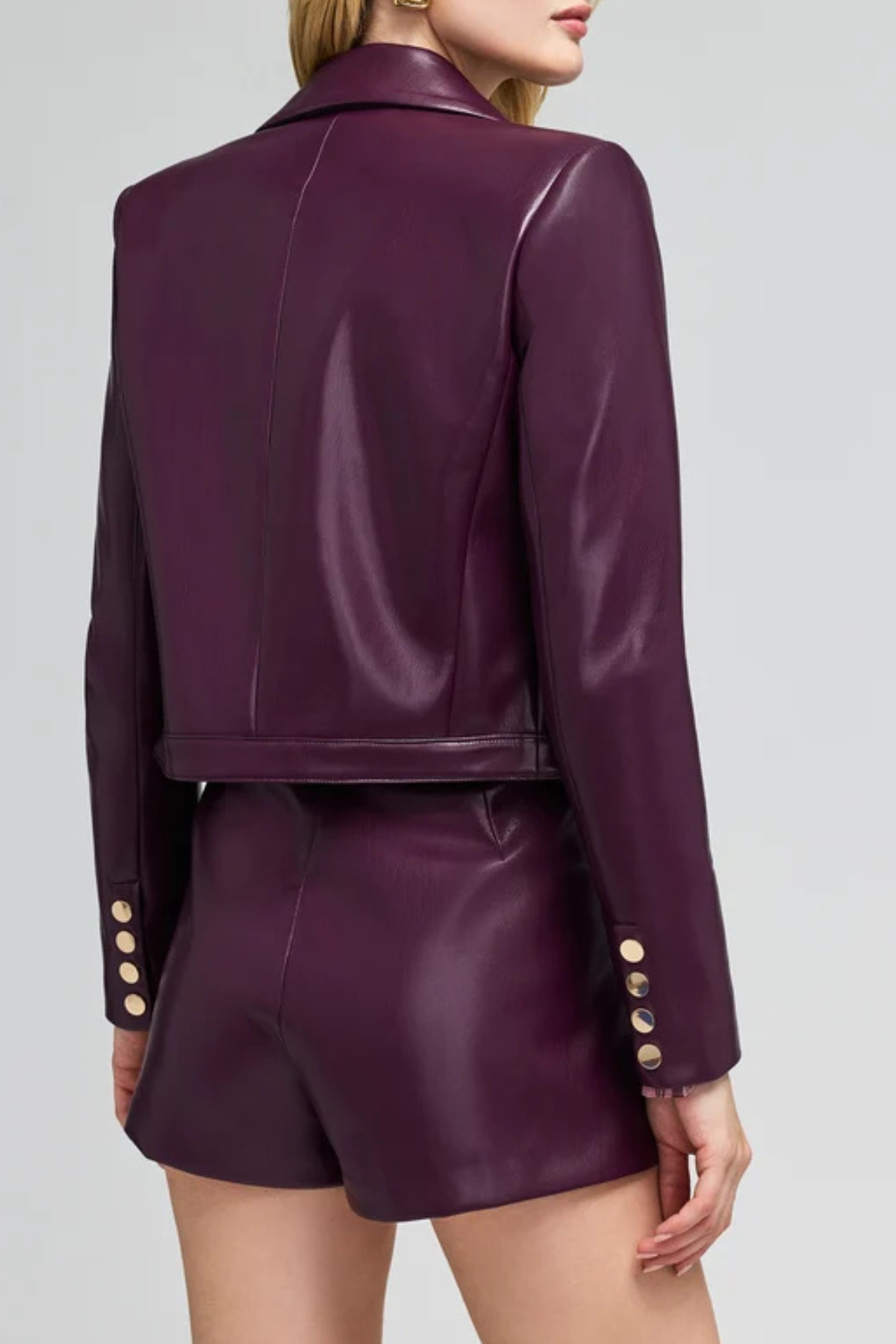 A model shown from the back wearing a matching deep purple faux-leather cropped blazer and shorts set, featuring gold-tone decorative buttons on the cuffs, against a light gray studio background.