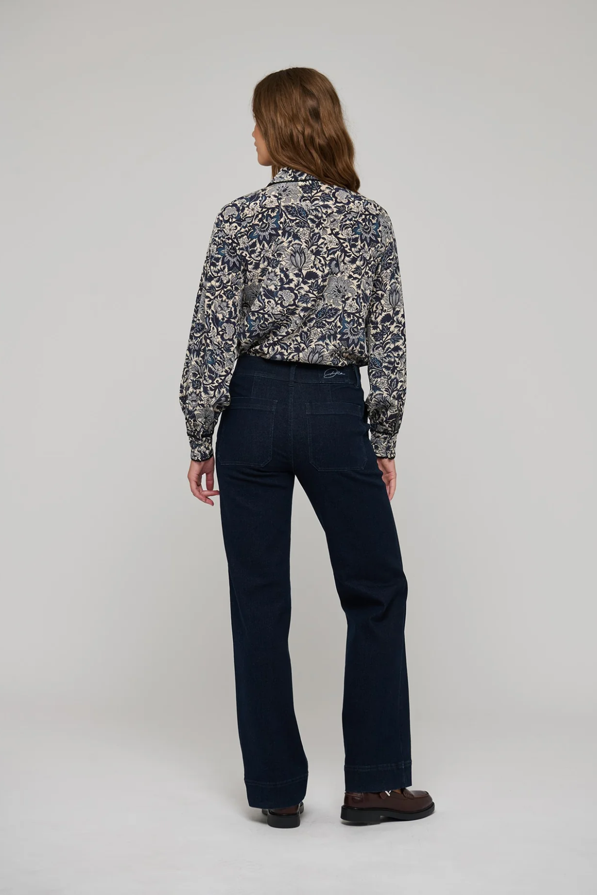 A model shown from the back wearing a long-sleeve button-up shirt with a blue, white, and gray floral pattern, paired with dark navy blue straight-leg jeans and brown shoes, against a light gray studio background.