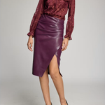 A model wearing a burgundy patterned long-sleeve blouse and a dark purple faux-leather wrap skirt with a side slit, paired with black pointed-toe heels, against a light gray studio background.