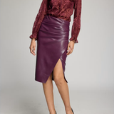 A model wearing a burgundy patterned long-sleeve blouse and a dark purple faux-leather wrap skirt with a side slit, paired with black pointed-toe heels, against a light gray studio background.