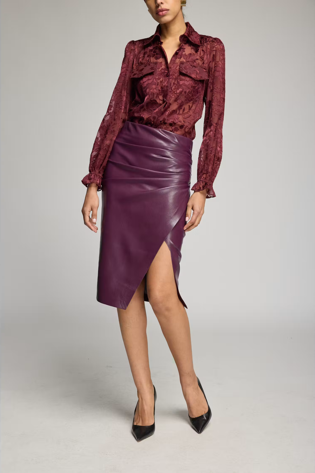 A model wearing a burgundy patterned long-sleeve blouse and a dark purple faux-leather wrap skirt with a side slit, paired with black pointed-toe heels, against a light gray studio background.