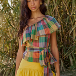 Woman wearing a colorful plaid top and yellow skirt standing in front of green foliage.