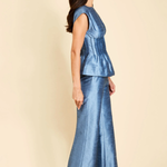 A woman modeling a matching two-piece blue satin set consisting of a sleeveless peplum top and wide-leg trousers, shown from a side profile against a solid beige background.