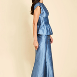 A woman modeling a matching two-piece blue satin set consisting of a sleeveless peplum top and wide-leg trousers, shown from a side profile against a solid beige background.