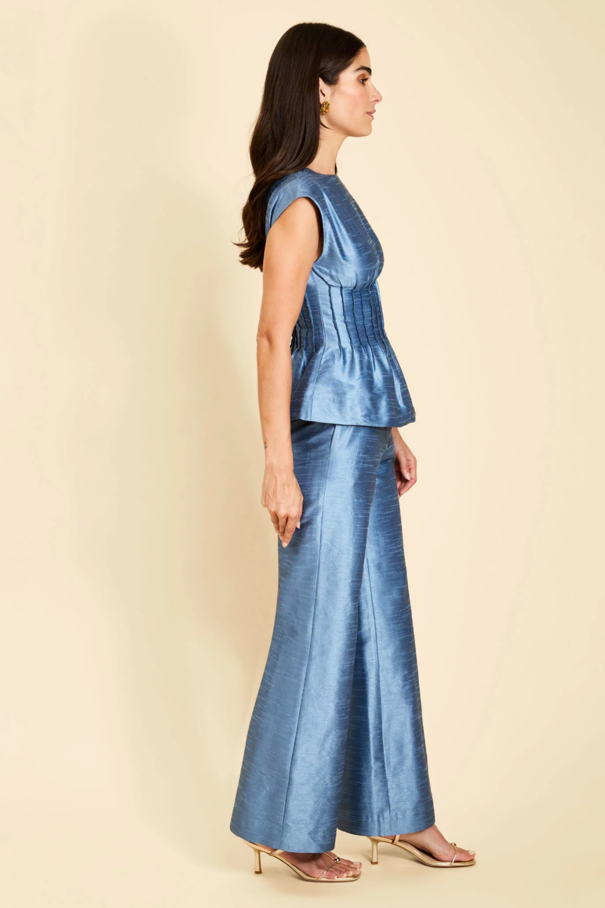 A woman modeling a matching two-piece blue satin set consisting of a sleeveless peplum top and wide-leg trousers, shown from a side profile against a solid beige background.