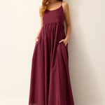 A woman modeling a sleeveless, smocked-bodice maxi dress in deep burgundy with side pockets, paired with dark brown strappy sandals, standing against a plain off-white wall.